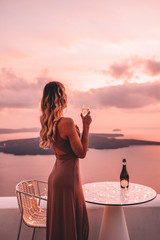 Young woman with blonde hair and purple dress watching the sunset in santorini greece - obrazy, fototapety, plakaty