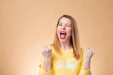 Happy successful young woman with raised hands shouting and celebrating success over yellow background