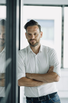 Portrait of confident mature businessman leaning against glass wall in office