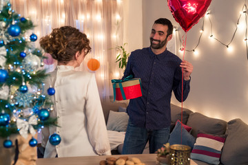 Handsome man surprising his girlfriend with a present and heart shaped balloon by the Christmas tree