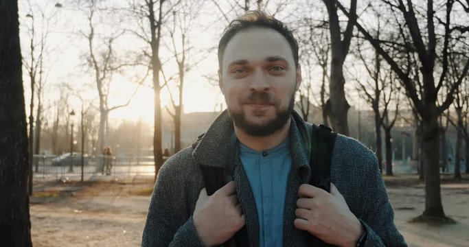 Cheerful handsome tourist man with camera smiling, walking backwards along warm autumn backlit park posing slow motion.