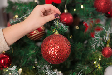 Female hand holds red ball on Christmas tree in artificial frost. In background decorated Christmas tree with big yellow bokeh. Fairytale atmosphere, idea house for new year. Closeup, selective focus.
