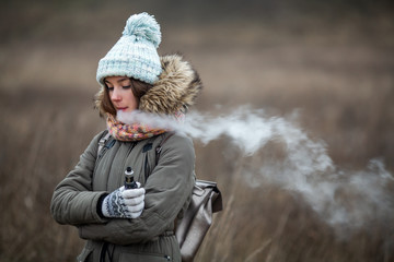 Vaping teenager. Young beautiful white teeage girl in casual clothes smokes an electronic cigarette on the street in the autumn. Electronic cigarette. Deadly bad habit.