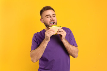 Young man eating tasty sandwich on yellow background