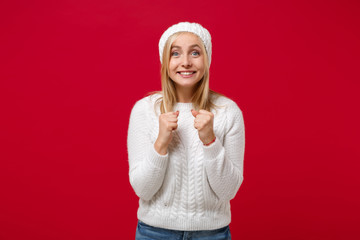 Excited young woman in white sweater hat isolated on red wall background studio portrait. Healthy fashion lifestyle cold season concept. Mock up copy space. Clenching fists waiting for special moment.