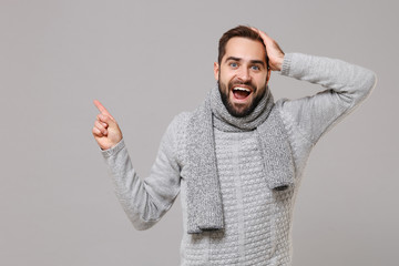 Fototapeta premium Excited young man in gray sweater, scarf posing isolated on grey background in studio. Healthy fashion lifestyle cold season concept. Mock up copy space. Pointing index finger aside put hand on head.