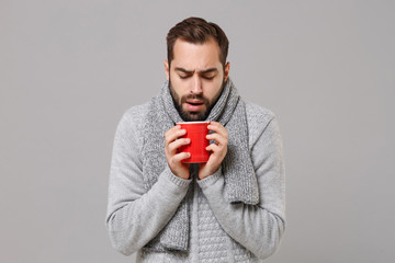 Frozen young man in gray sweater, scarf posing isolated on grey wall background, studio portrait. Healthy fashion lifestyle, cold season concept. Mock up copy space. Holding red cup of tea or coffee.