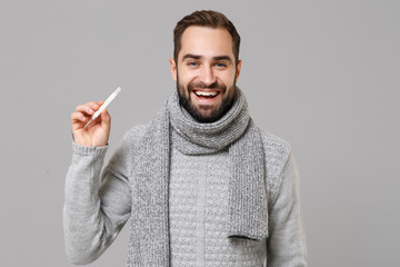 Smiling young man in gray sweater, scarf isolated on grey wall background studio portrait. Healthy lifestyle, ill sick disease treatment, cold season concept. Mock up copy space. Hold thermometer.