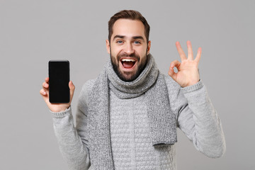 Man in gray sweater, scarf isolated on grey background. Healthy fashion lifestyle, people emotions, cold season concept. Mock up copy space. Hold mobile phone with empty screen, showing OK gesture.