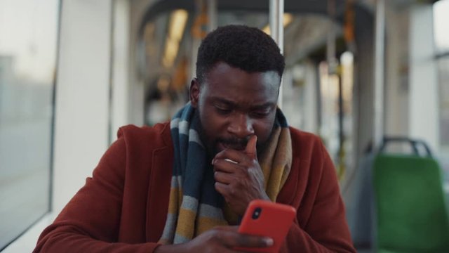 Sick poor handsome man sneezing allergy getting cold driving in tram to work. Modern young black businessman using smartphone reading news traveling in public transport.