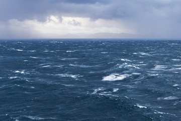 Grobe See im Atlantik, Europäisches Nordmeer vor Norwegen, mit Wellen und Gischt bei stürmischem Wind. Am Horizont dunkle Wolken mit Auflockerung, durch die die Küste zu sehen ist.