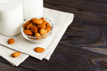 Close-up of almond milk in a glass mug and a refillable glass bottle, next to it lie almonds and a textile napkin on a wooden table.