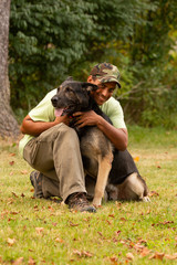 A man kneels down and hugs his german shepherd dog on a grassy lawn with trees in the background. 