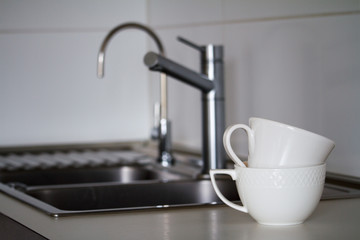 Two clean white tea cups on the background of a metal sink with a water tap, faucet and drinking tap, selective focus