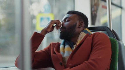 Allergic sick african man sneezing into paper tissue coughing and suffering bad virus of flu driving in public transport. Unwell young businessman going home after work.