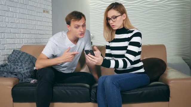 Excited couple finding amazing smartphone content sitting on a couch in the living room at home. Young couple wins a lottery or stakes