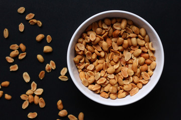 Peanuts in a white bowl as seen from above with some peanuts spreaded over a dark background