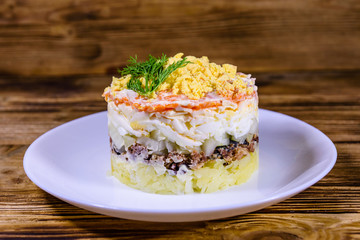 Plate with traditional russian salad Mimosa on a wooden table. Layered salad with potatoes, sardine, cheese, carrot, eggs and mayonnaise