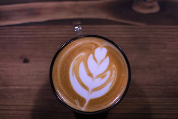 A cup of coffee with leaf pattern in a glass cup on wooden background.