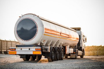 Truck with trailer, tank with flammable liquid, under a large bridge at the pier on the river bank, sunset light, white and red cars standing on gravel