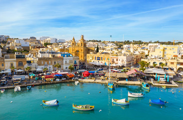 Fototapeta na wymiar Landscape view of fishing village Marsaxlokk. Traditional maltese boats on the sea, main church, coastline, blue sly. Malta