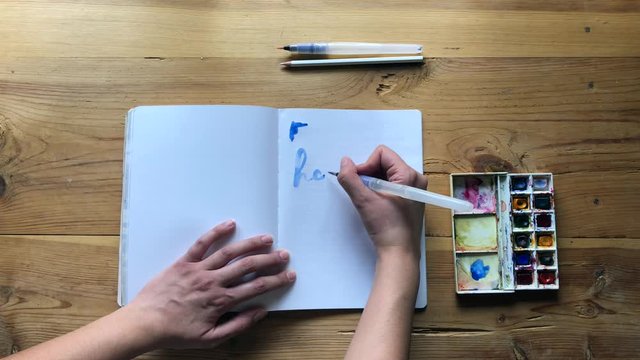 Young Woman artist draw with watercolor paints with a brush in a bullet journal pages. Blue watercolor painter on white canvas. Woman hands painting a christmas message. Slow motion.