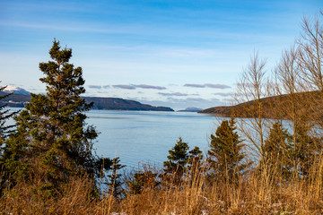 Seascape of a calm ocean, blue sky, clouds, mountains and trees in the foreground.