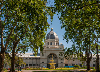 Fototapeta premium Melbourne, Australia - November 18, 2009: Pagoda-style fountain with statues in front of Royal Exhibition Building, central part with dome, under blue sksy. Green foliage of Carlton Gardens.