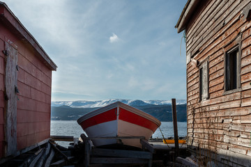 A small red and white open boat pulled up on land between two red worn buildings. There is a cloudy sky and snow covered mountains in the background.