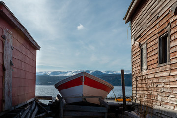 A small red and white open boat pulled up on land between two red worn buildings. There is a cloudy sky and snow covered mountains in the background.