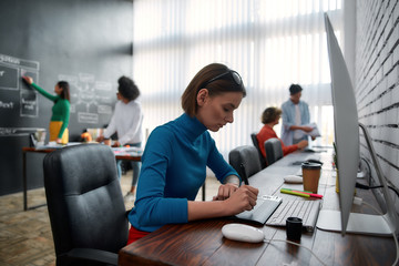 Professional graphic designer. Side view of young creative woman using digital graphics tablet while sitting at her workplace in the creative agency. Web designer is drawing on tablet with pen stylus