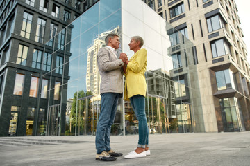 Fototapeta na wymiar I wish to be with you forever. Full-length of elegant and stylish mature couple holding hands and looking at each other while standing against modern glass building