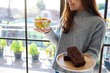 A woman holding and choosing food to eat between a plate of brownie cake and a bowl of vegetables salad