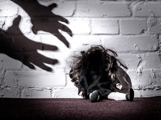 A doll sits on the floor and leans against a wall. The shadows of hands reach for her. Concept:...