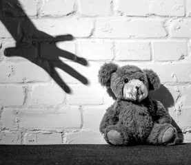 A teddy is sitting on the floor and leaning against a wall. The shadow of a hand reaches for it....