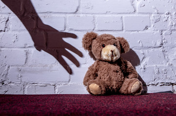 A teddy is sitting on the floor and leaning against a wall. The shadow of a hand reaches for it....
