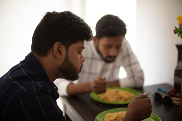 Two young tall, dark and handsome Indian Bengali men in casual wear having lunch together sitting in a dining table in white background. Indian lifestyle and fashion.
