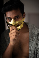 An young,tall,dark and handsome Indian Bengali man wearing a golden mask while standing in front of a window in studio background. Indian lifestyle.