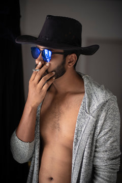 An young,tall,dark and handsome Indian Bengali man wearing a hat,golden mask and cigarette in hand standing in front of a window in studio background. Indian lifestyle.