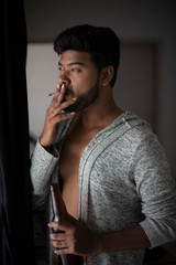 An young tall, dark and handsome Indian Bengali man in a front open western jacket smoking while looking outside standing in front of a window in studio background. Indian lifestyle and fashion.