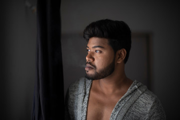 An young tall, dark and handsome Indian Bengali man in a front open western jacket smoking while looking outside standing in front of a window in studio background. Indian lifestyle and fashion.