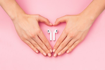 Heart with female hands holding wireless earphones on pink background. Young woman hands in shape of heart and earbuds on flatlay.