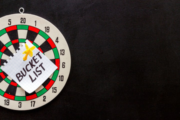 Obraz na płótnie Canvas Bucket list attached by dart to dart board on black background top-down copy space