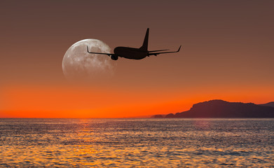 Airplane flying above tropical sea at sunset with full moon 