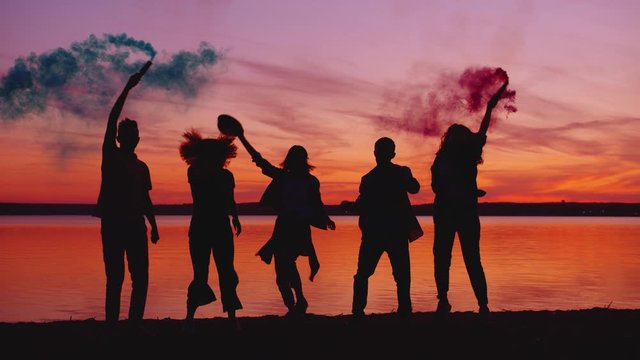 People men and women are dancing on the beach at sunset holding smoke flares enjoying outdoor party together. Entertainment, youth and lifestyle concept.