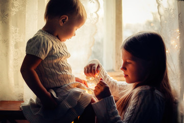 children in cozy dresses gently play, window