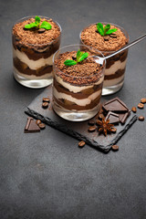 Classic tiramisu dessert in a glass cup and pieces of chocolate on stone cutting board on dark concrete background