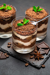 Classic tiramisu dessert in a glass cup and pieces of chocolate on stone cutting board on dark concrete background