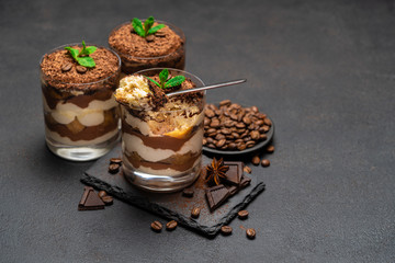 Classic tiramisu dessert in a glass cup and pieces of chocolate on stone cutting board on dark concrete background