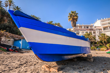 Blue white wooden boat on the beach of the Spanish Mediterranean coast, Costa del Sol. Sandy beach by day in sunlight with palm trees and buildings in the background